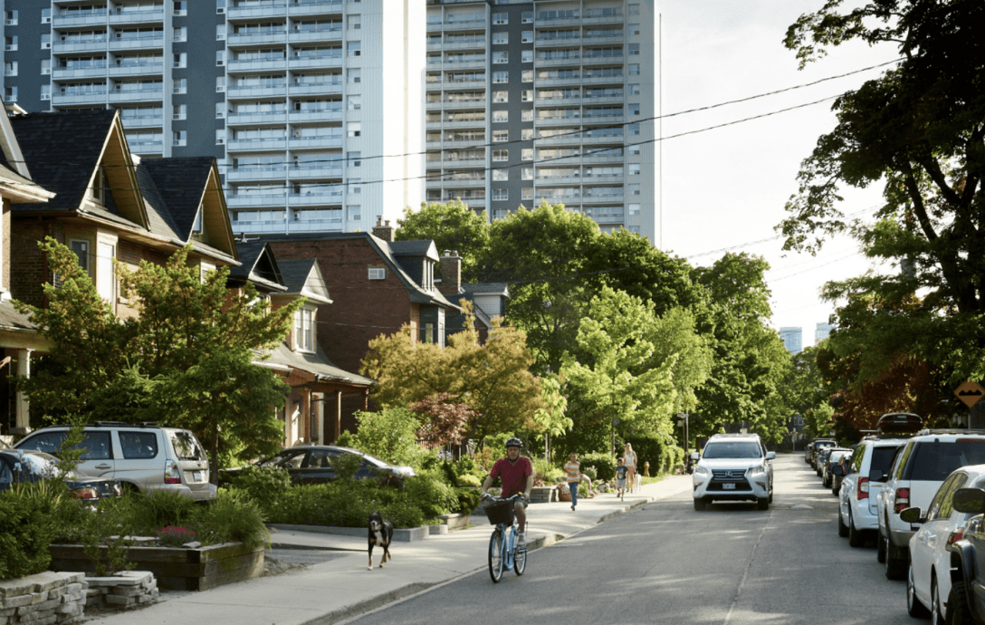 A cyclist riding along a residential street with a dog and family on the sidewalk and apartment buildings in the backdrop.