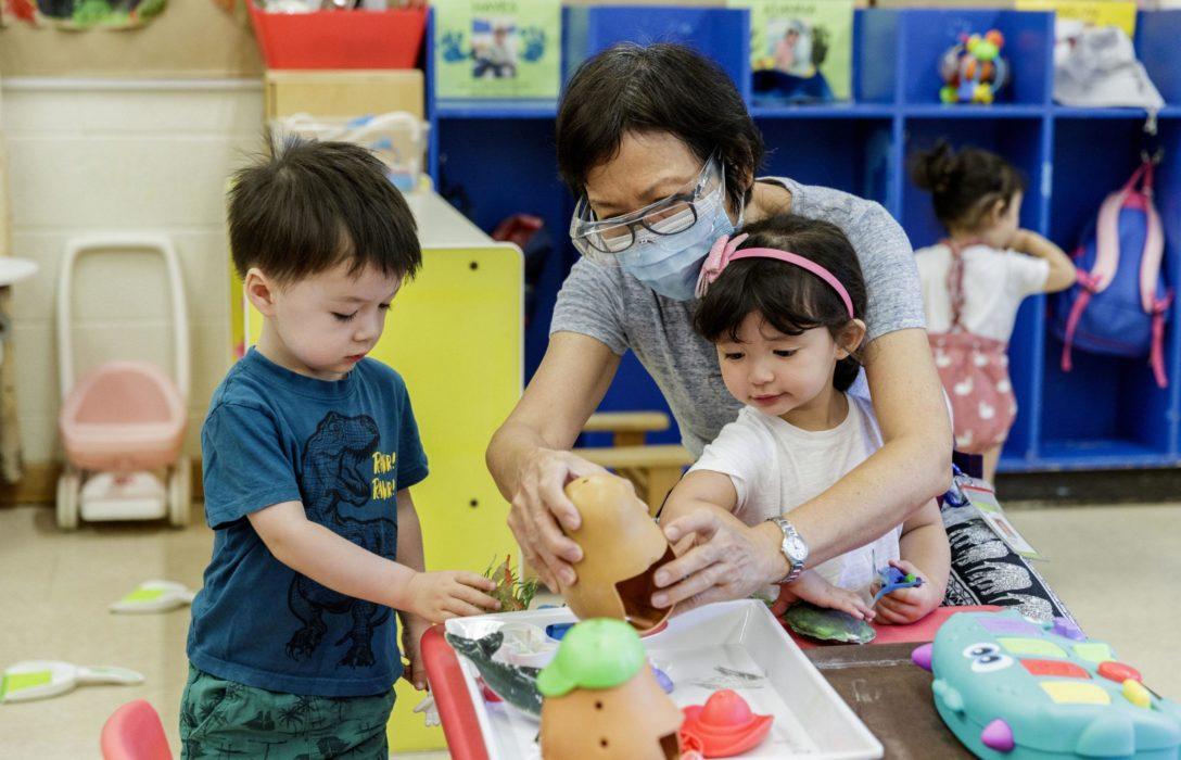 Educator wearing a mask and playing at a table with two young children in a classroom environment.