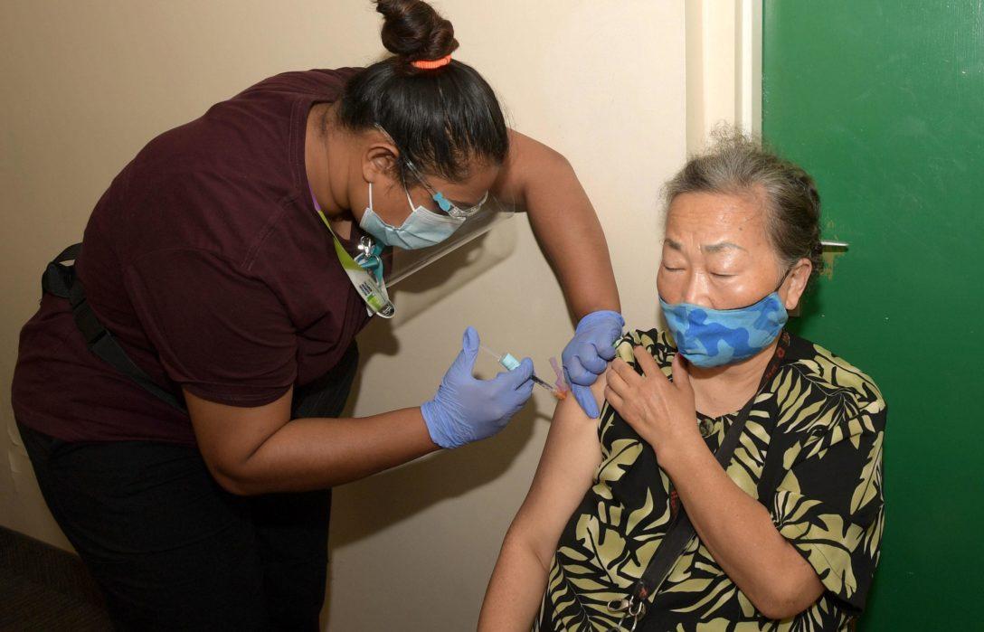 A senior sitting in an apartment hallway wearing a mask, receiving a vaccination from WoodGreen staff.