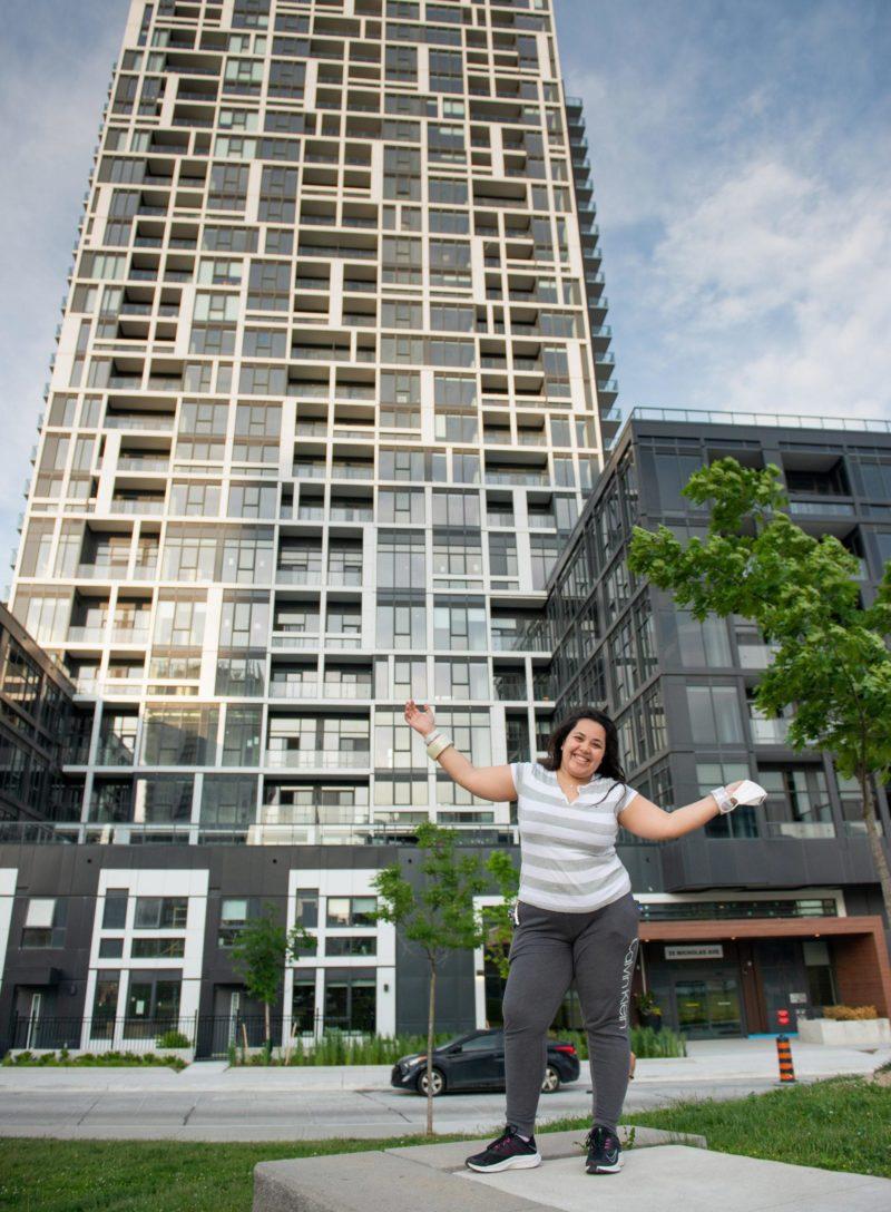 A woman posing outside an apartment building with her arms outstretched and proudly smiling.