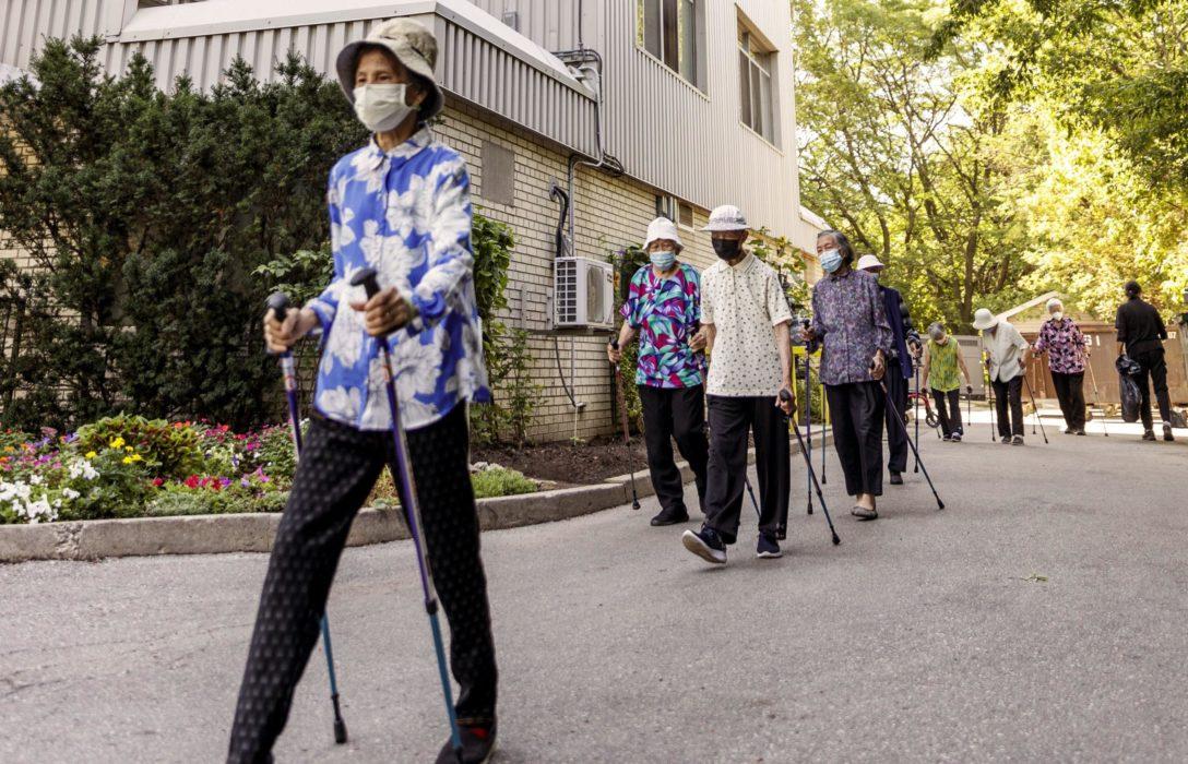 A group of seniors walking together alongside a building wearing masks and using walking poles.
